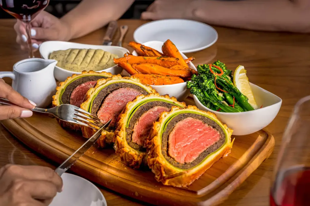 A wooden board featuring sliced Beef Wellington with a golden pastry crust, served with roasted carrots and broccolini.