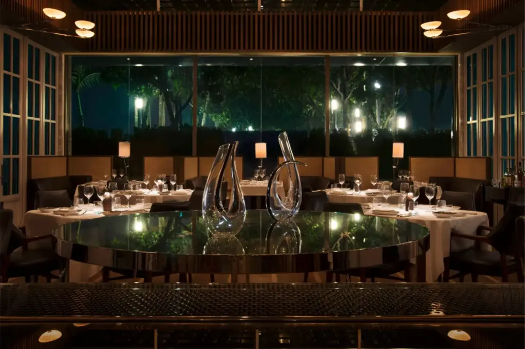 An eye-level, medium shot captures the sophisticated and dimly lit interior of an upscale restaurant at night, representing the file "8-Where to Dine at Marina Bay Sands.webp". In the foreground, two sculptural, clear glass wine decanters stand on a large, reflective circular table with a polished metallic rim, mirroring the ambient light. Beyond this central feature, the main dining room is arranged with tables dressed in white tablecloths, set with crystal glassware, white plates, and individual small lamps that cast a soft glow. The back of the restaurant is dominated by an expansive floor-to-ceiling glass window, revealing a view of illuminated green trees and outdoor lighting against the dark night sky, adding a serene and luxurious backdrop to the refined dining space.