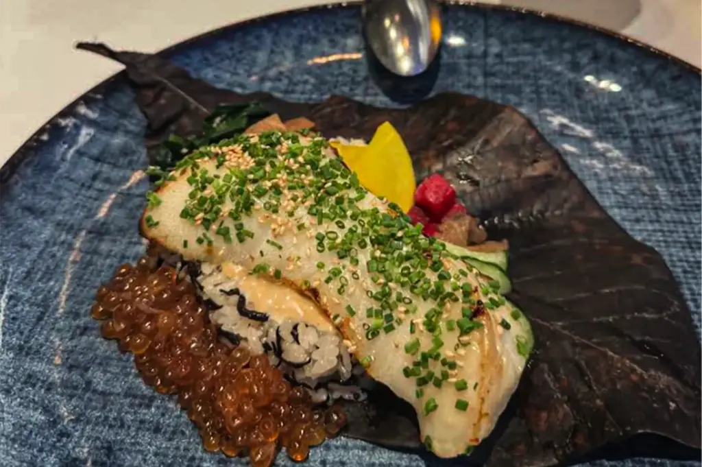 A close-up, high-angle shot captures an exquisitely plated seafood dish presented on a textured, dark blue ceramic plate, representing the file "9-Where to Dine at Marina Bay Sands.webp". The meal is artfully arranged over a large, dried brown leaf, featuring a perfectly cooked fillet of white fish heavily garnished with finely chopped green chives and a dusting of sesame seeds. The fish rests on a bed of seasoned white rice flecked with dark seaweed, accompanied by a generous cluster of translucent, orange ikura (salmon roe) pooling on the left side. A vibrant assortment of pickled components garnishes the top right, including bright yellow takuan radish, a deep red beet cube, and thinly sliced cucumbers, creating a visually striking and sophisticated culinary presentation.