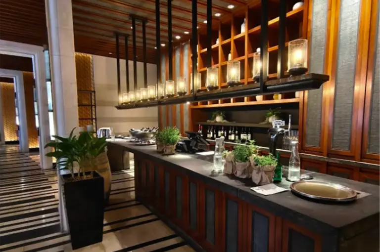 An elegant restaurant interior featuring a long dark counter decorated with small potted plants, glass bottles, and serving trays. Warm pendant lights hang above the counter, while wooden shelves and soft ambient lighting create a sophisticated and cozy atmosphere with modern Asian-inspired design elements.