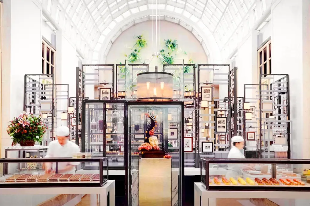 Two pastry chefs in white uniforms preparing desserts behind glass display cases at Colony restaurant in Singapore, featuring high arched ceilings and elegant modern decor.
