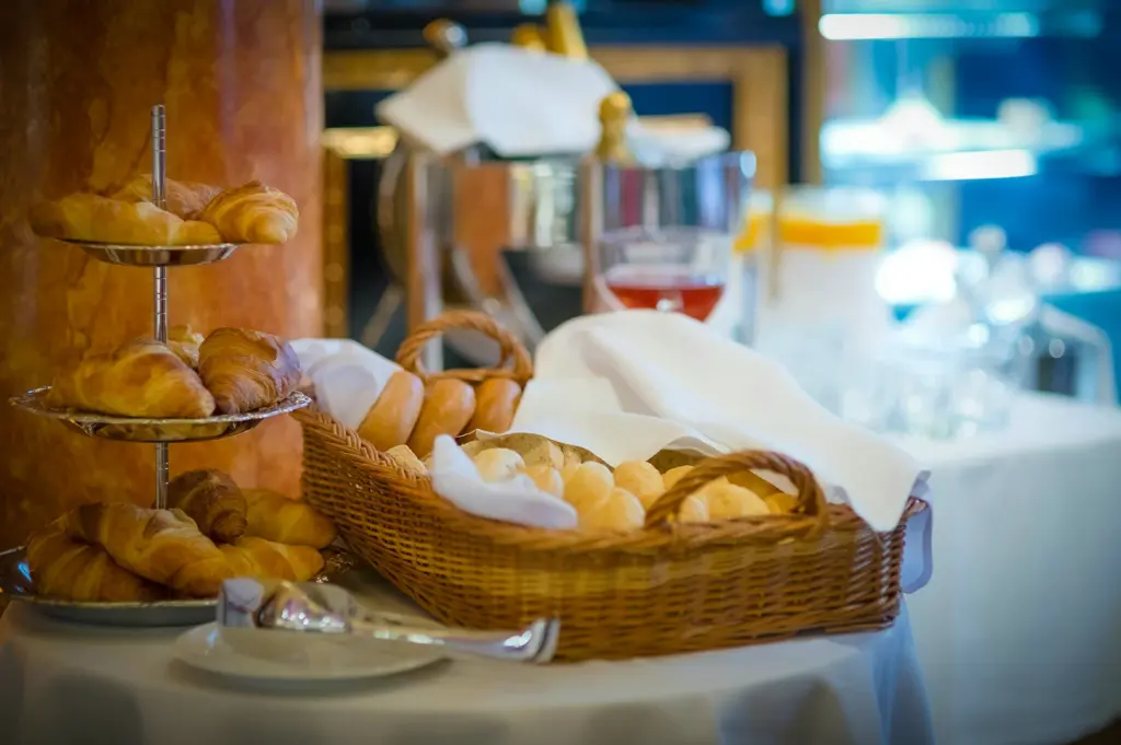 Fresh croissants, bread rolls and pastries displayed in baskets and serving trays at an elegant hotel breakfast buffet setup with warm ambient lighting