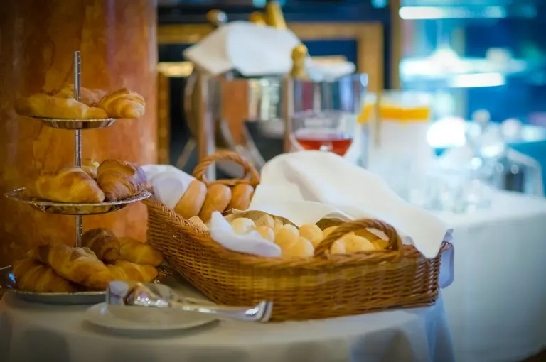 Fresh croissants, bread rolls and pastries displayed in baskets and serving trays at an elegant hotel breakfast buffet setup with warm ambient lighting