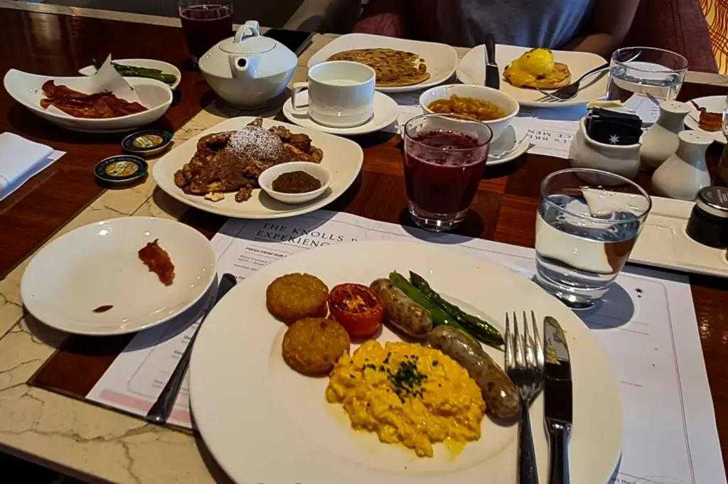 A breakfast spread arranged on a wooden restaurant table, featuring scrambled eggs, sausages, roasted tomato, asparagus, and hash browns on a large white plate in the foreground. Surrounding dishes include waffles or pancakes, bacon, pastries, tea, juice, and coffee, creating a cozy and abundant brunch setting with drinks and condiments placed around the table.