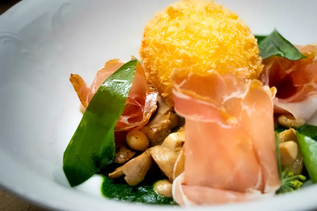 Gourmet dish featuring a golden crispy breaded cheese ball served with prosciutto slices, sautéed mushrooms, and greens on a white plate.