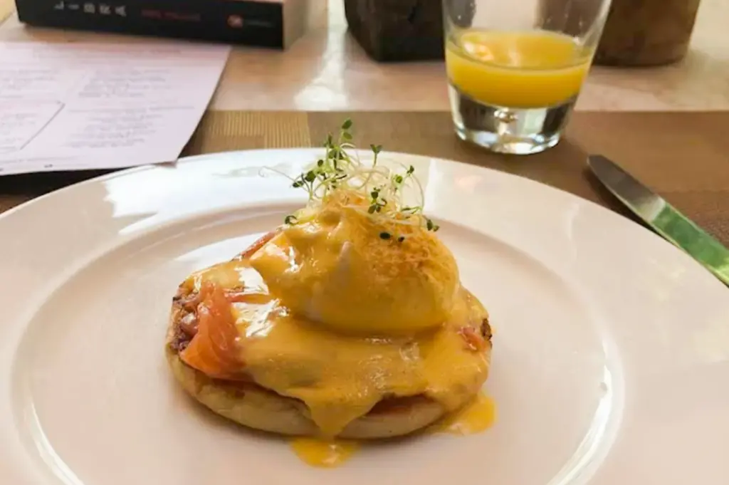A close-up of an eggs Benedict-style breakfast dish served on a white plate, featuring a poached egg topped with creamy hollandaise sauce over smoked salmon and an English muffin. Fresh microgreens garnish the top, while a glass of orange juice and dining utensils sit in the softly blurred background.