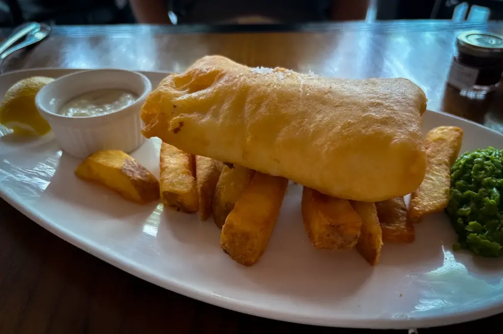 Fish and chips at Bread Street Kitchen Singapore featuring crispy battered fish, thick-cut fries, mushy peas, tartar sauce, and lemon wedge on a white plate
