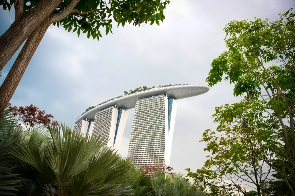 Low-angle view of Marina Bay Sands in Singapore framed by lush green trees, showing the iconic rooftop SkyPark under a cloudy sky.
