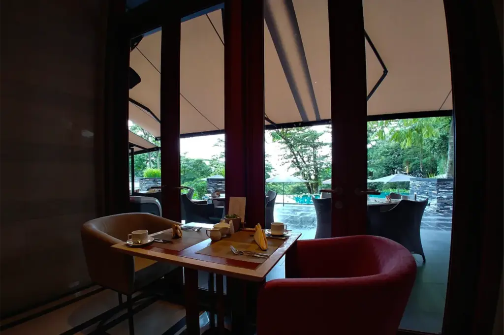 A cozy restaurant dining area with a small table set for two beside tall glass doors overlooking an outdoor patio and lush greenery. Warm indoor lighting contrasts with the bright natural view outside, while cushioned chairs and neatly arranged tableware create an intimate and elegant atmosphere.