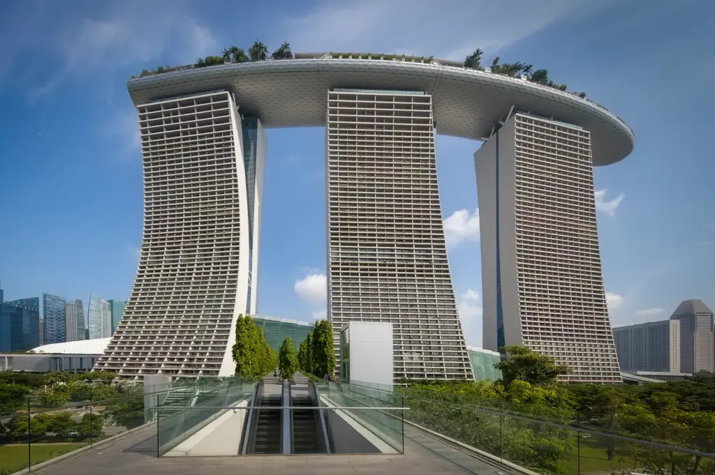 Frontal view of Marina Bay Sands in Singapore with its three towers and SkyPark rooftop, seen from a walkway with greenery and city skyline in the background.