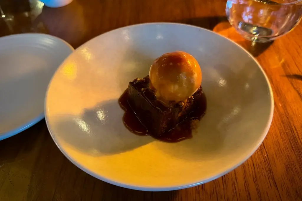 A dessert plate with a brownie topped with a scoop of ice cream, drizzled with caramel sauce, sits on a wooden table. The setting is warmly lit.