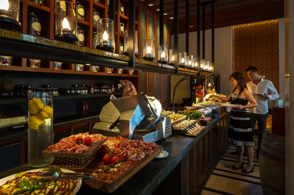 A warmly lit buffet restaurant interior featuring a long serving counter lined with cured meats, fresh dishes, and buffet selections. Decorative pendant lights hang above dark wood shelving, while two diners serve themselves food, creating an elegant and upscale dining atmosphere.