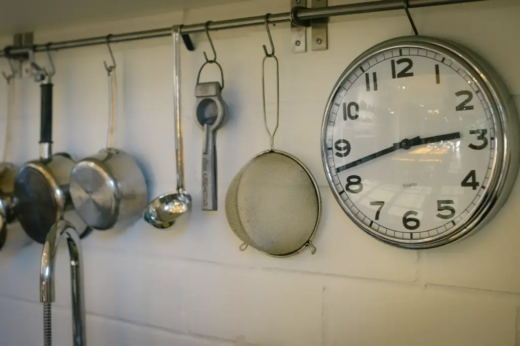 Kitchen wall with hanging metal utensils including ladle and strainer beside a round wall clock showing time in a bright cooking space.