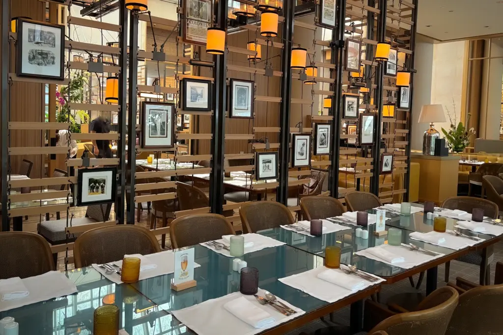 Elegant dining area at Colony Singapore featuring neatly set tables with white napkins, glass-topped tables, and a decorative partition wall with vintage framed photos and warm amber lighting.