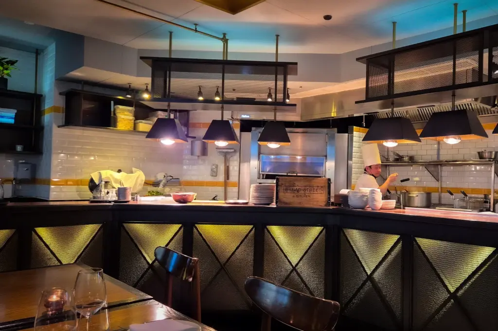 Interior of Bread Street Kitchen Singapore featuring an open kitchen concept, chefs preparing food, industrial-style lighting, and modern restaurant seating with warm ambient decor