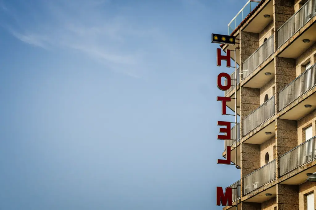 A low-angle, medium shot captures the side corner of a multi-story concrete hotel building against a vast blue sky, representing the file "1-Some Hotels Become Part.webp". The beige, textured stone building features a vertical stack of identical balconies with metallic railings and small circular windows on the side wall. Attached to the corner is a large, vertical, three-dimensional sign spelling out "HOTEL" in bold red block letters, with the letter "M" partially visible at the very bottom. Above the lettering, a small black rectangular sign displays three yellow stars, indicating the hotel's rating. The minimalist composition places the building on the right third of the frame, leaving the remaining left side dedicated to a clear, pale blue sky with faint, wispy clouds.