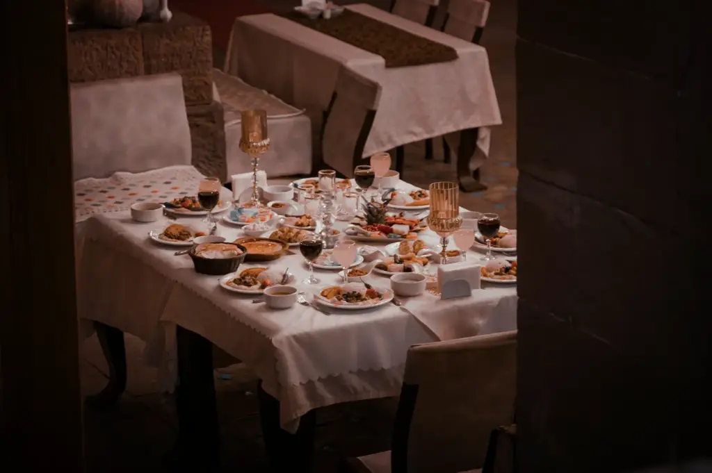 A high-angle, medium shot captures an elegant and sumptuously laid dining table in a dimly lit, sophisticated restaurant, representing the file "1-Some Meals Become Attached to a Place Forever.webp". The long table is draped in a crisp white scalloped tablecloth and surrounded by cushioned chairs, heavily laden with a diverse feast of individual plates of meat and rice, small shared bowls, appetizers, a basket of bread, and multiple glasses filled with red wine and light-colored beverages. Two tall, decorative gold chalice-style candle holders stand symmetrically on the table, adding a touch of grandeur to the setup. In the softly blurred background, another impeccably set dining table sits empty, while a dark stone wall partially frames the right foreground, enhancing the intimate, moody ambiance of the dining space.