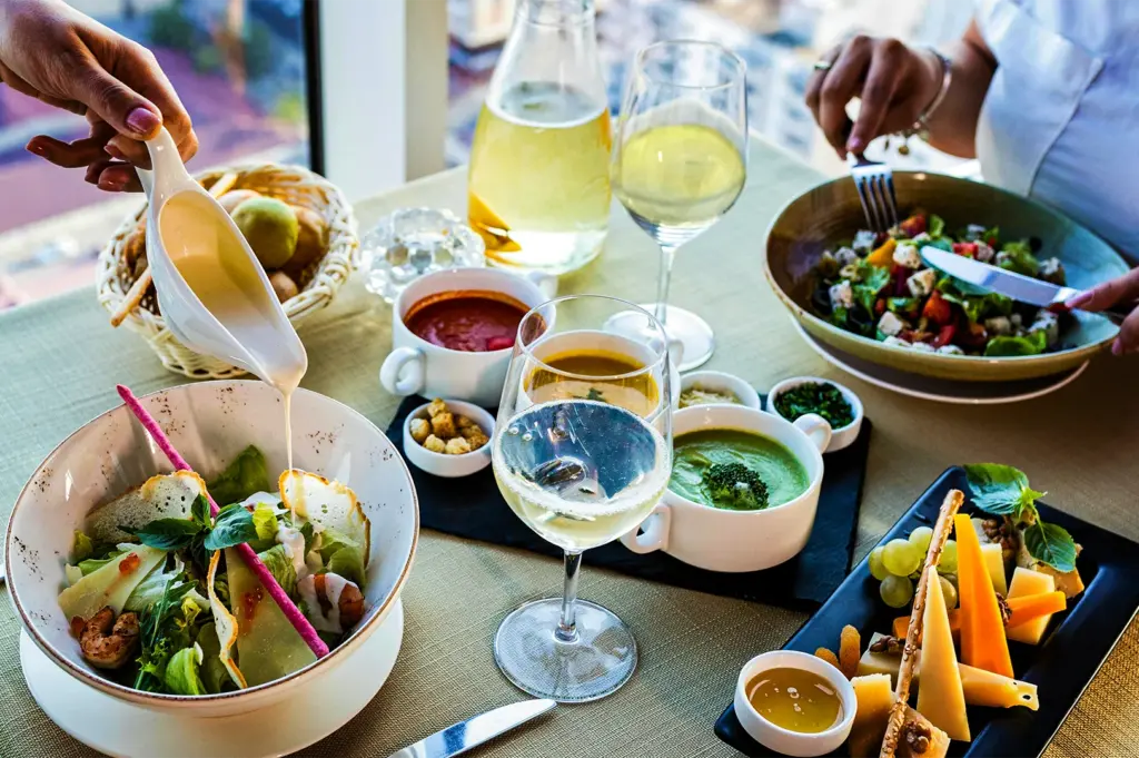 A high-angle, medium shot captures an elegant and abundant lunch spread served at a high-rise restaurant table, representing the file "1-Some Restaurants Turn Dinner Into Memory_2.jpg". On the left, a person's hand pours a creamy white dressing from a porcelain boat onto a gourmet salad filled with fresh greens, grilled shrimp, and shaved parmesan cheese. The center of the table features a flight of three vibrant soups—tomato red, butternut squash orange, and broccoli green—presented in small white bowls alongside croutons and herbs on a black slate. In the foreground, a crystal wine glass filled with white wine stands next to a rectangular black platter of assorted cheeses, grapes, nuts, and a small dish of honey, while a second diner enjoys a large Mediterranean salad in the background against a bright window view overlooking a city skyline.