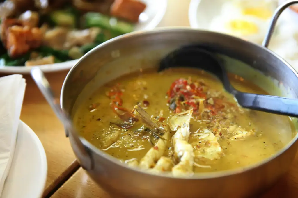 A close-up, slightly high-angle shot captures a comforting, aromatic bowl of soup curry served on a wooden table, representing the file "1-Soup Curry The Bowl.webp". A deep stainless steel pot with small side handles contains a rich, yellowish-green curry broth filled with tender chicken, visible bones, and soft, stewed ingredients garnished with red chili flakes. A black plastic serving spoon rests inside the pot, dipping into the savory soup. In the blurred background, other dining plates are partially visible, including a colorful stir-fry with green vegetables on the left and sliced hard-boiled eggs on the right, emphasizing a hearty and flavorful casual meal setting.