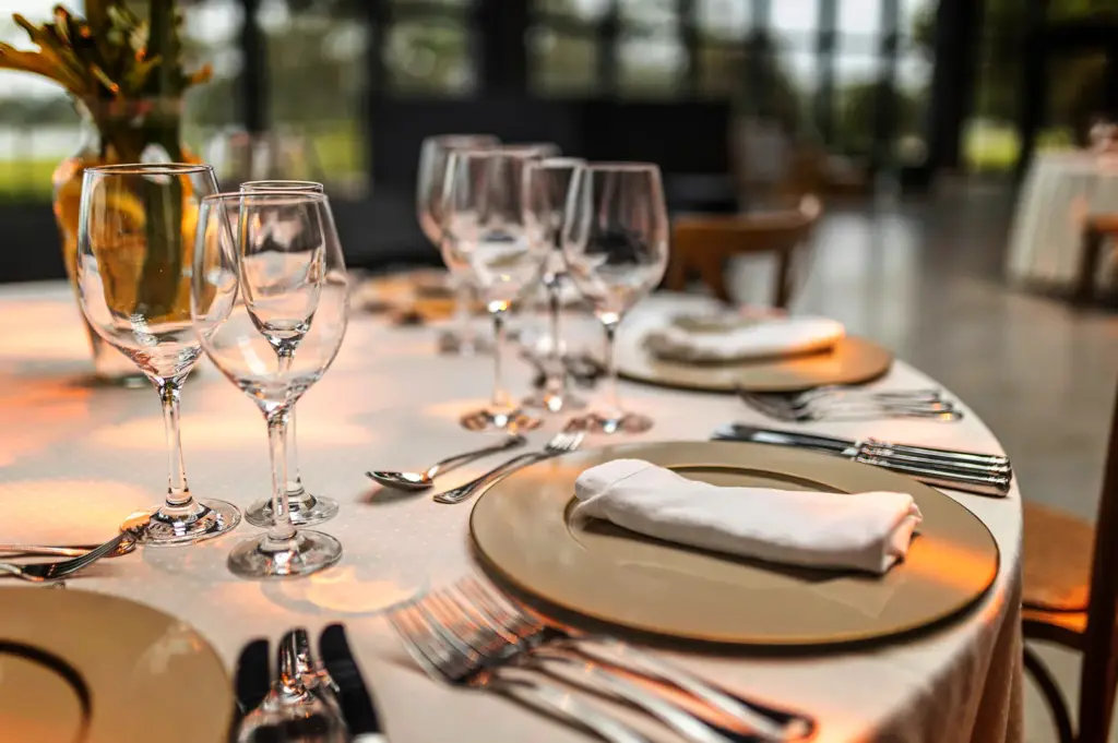 A close-up, shallow-depth-of-field shot captures an elegantly set round dining table inside a modern, spacious restaurant, representing the file "1-The Best Fine Dining Experiences Never Feel Forced.webp". In the foreground, a pristine white tablecloth is meticulously arranged with smooth, beige chargers holding neatly rolled white cloth napkins, flanked by polished silver forks, knives, and spoons. Clusters of delicate, empty crystal wine glasses stand gleaming on the table, beautifully catching the warm, golden lighting that reflects off the surfaces. The background softly blurs out, revealing glimpses of wooden chairs, expansive floor-to-ceiling windows, and a sophisticated, airy dining room ambiance.