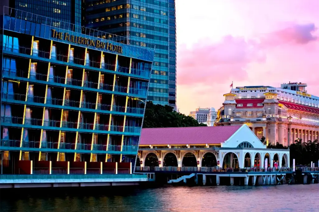 An eye-level, wide shot captures a striking architectural contrast along a waterfront at sunset, representing the file "1-The Fullerton Bay Hotel.webp". On the left, the modern, diamond-faceted glass facade of The Fullerton Bay Hotel rises from the water, its rows of balconies glowing with warm interior yellow lights that contrast beautifully with the blue glass. Next to it sits a low-rise, historic white building with a red roof and a series of elegant arches, built over a pier structure on the water. In the background, the grand, neoclassical facade of The Fullerton Hotel stands beneath a vibrant sky filled with soft pink, purple, and orange clouds, while the calm water in the foreground beautifully catches the reflection of the evening light.