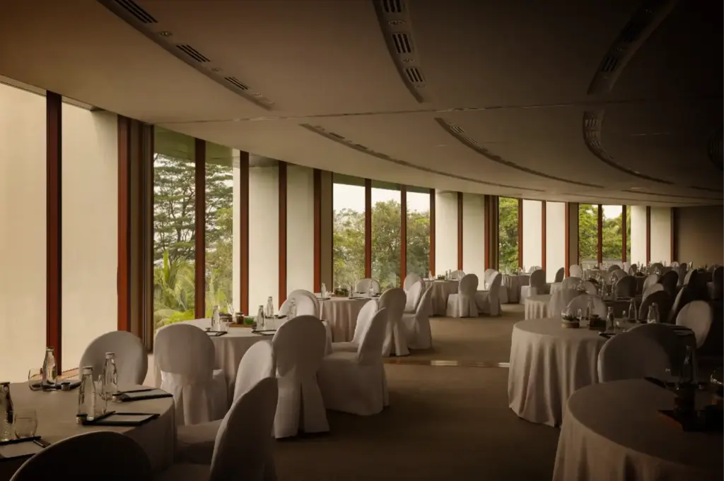 An eye-level, wide shot captures a large, upscale banquet hall or dining room with a curved wall of floor-to-ceiling windows, representing the file "1-Where Stillness Meets Luxury.webp". The spacious room features numerous round dining tables meticulously set with white tablecloths, glassware, and bottled water, surrounded by chairs draped in elegant white fabric covers. The sweeping, segmented glass facade bathes the carpeted space in muted natural light while offering a serene view of lush green trees and tropical foliage outside. The curving architecture of the ceiling lines mirrors the wall of windows, creating a sophisticated, tranquil, and luxurious event setting.