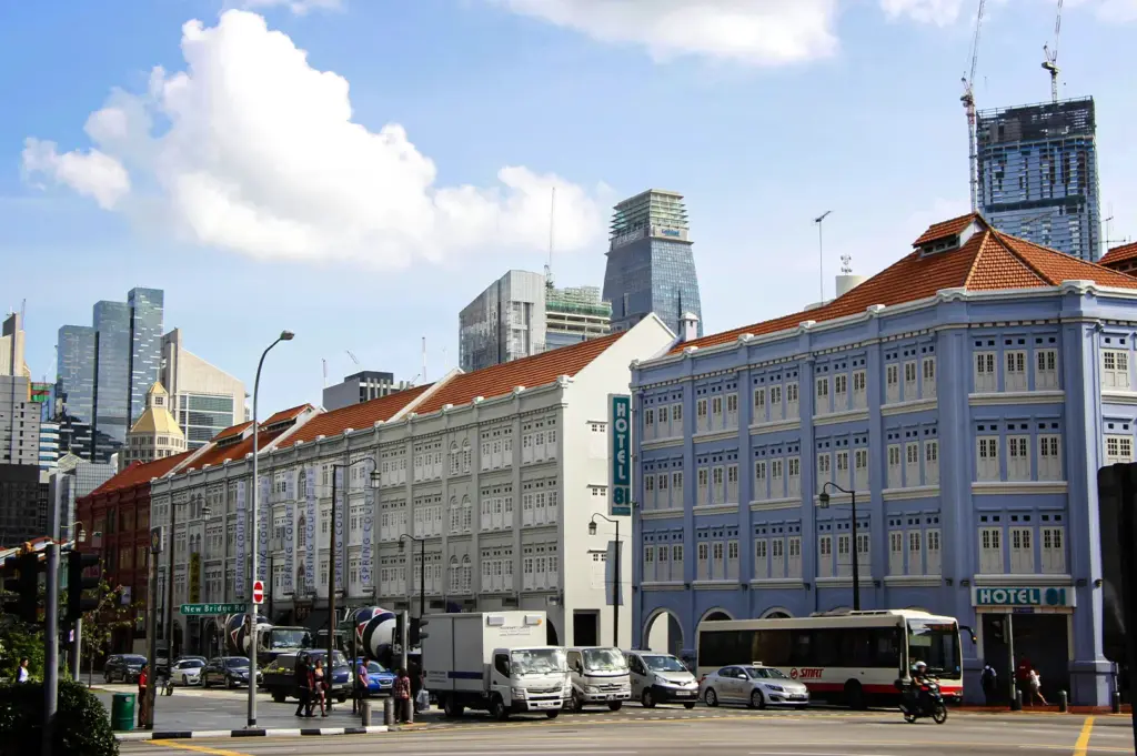 An eye-level, wide shot captures a bustling city intersection where heritage architecture meets a modern skyline, representing the file "1-Why the Right Hotel Can Change.webp". In the foreground, a multi-story colonial-style shophouse building stretches across the scene, featuring a distinct terracotta-tiled roof and painted in segments of white and a soft periwinkle blue, with a vertical sign reading "HOTEL 81" prominently displayed. Traffic, including white delivery trucks, a transit bus, a motorcycle, and several cars, navigates the wide asphalt junction of New Bridge Road under a bright blue sky filled with fluffy white clouds. In the background, sleek, glass-faced skyscrapers and a construction crane tower over the historic buildings, creating a striking visual contrast of old and new urban development.
