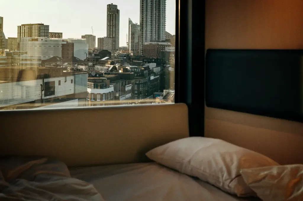 An eye-level, medium shot looks out from a modern, minimalist hotel room onto a dense city skyline, representing the file "2-Some Hotels Become Part.webp". In the dim foreground, an unmade bed features a white pillow resting on white sheets next to a beige padded headboard panel that extends along the wall under a large horizontal window. Through the glass, a sprawling urban landscape unfolds under a pale, hazy sky, showing a mixture of shorter brick buildings, low-rise apartment blocks, and several sleek, towering skyscrapers rising in the distance, juxtaposing a cozy private interior with the grand scale of the city outside.