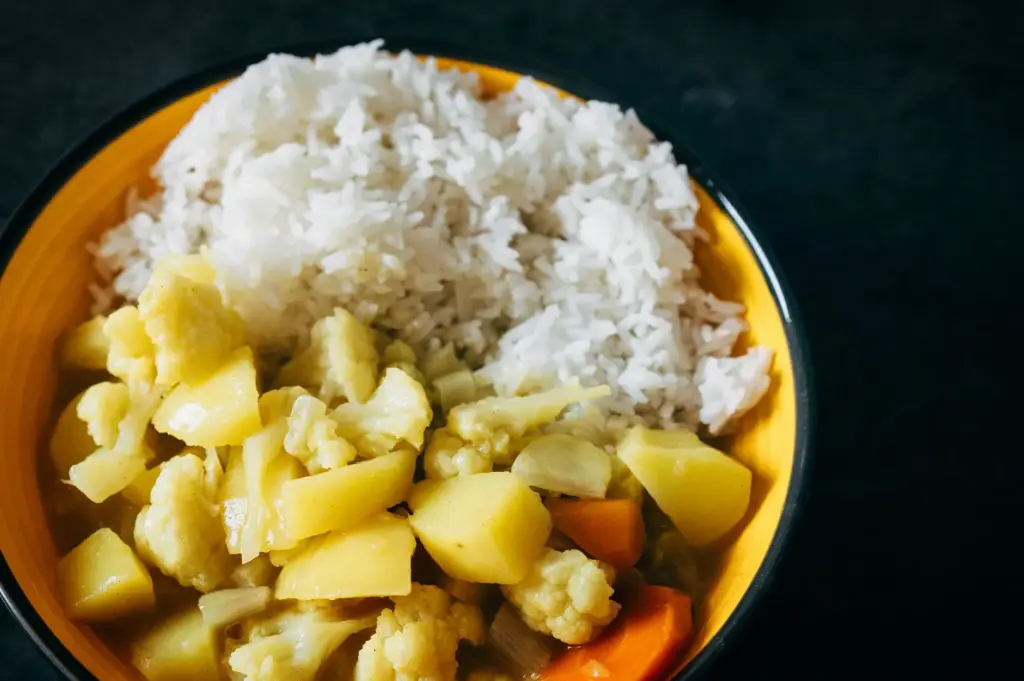 A high-angle, close-up shot captures a simple and hearty vegetable curry served in a vibrant yellow ceramic bowl, representing the file "2-Soup Curry The Bowl.webp". The bowl is cleanly split into two halves: the top half is filled with a generous mound of fluffy, white jasmine rice, while the bottom half contains a thick, yellowish curry packed with chunks of tender potatoes, cauliflower florets, onions, and bright orange carrots. The dish sits against a solid, dark black background, highlighting the bright colors of the ingredients and providing a clean, appetizing look at the homestyle meal.