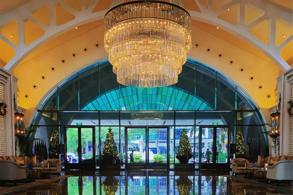 A symmetrical, eye-level shot captures the grand, luxurious lobby entryway of a building, representing the file "2-The Fullerton Bay Hotel.webp". Hanging prominently from the center of a massive, cream-colored vaulted ceiling with geometric architectural cutouts is a colossal, multi-tiered crystal chandelier that sparkles with warm, golden light. Below it, a towering glass-walled facade with an arched frame looks out onto a driveway area, where illuminated green Christmas trees stand symmetrically just inside the glass doors. The highly polished, dark marble flooring in the foreground acts as a perfect mirror, casting a vivid reflection of the entire majestic entryway, the twinkling trees, and the opulent seating lounges tucked neatly along both sides of the frame.