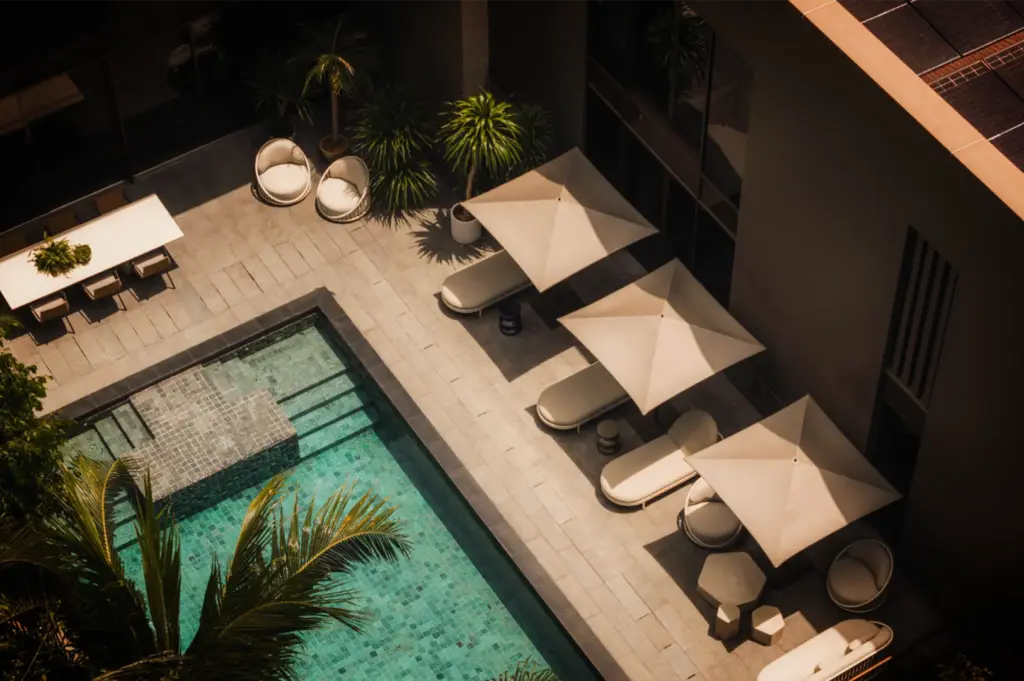 A high-angle, birds-eye view captures a serene and luxurious outdoor patio and swimming pool area, representing the file "2-Where Stillness Meets Luxury.webp". On the left, a rectangular pool with turquoise water and tiled steps is framed by a palm tree in the lower corner. The stone-tiled pool deck features an organized row of lounge chairs shaded by large, square beige umbrellas alongside various modern, rounded patio chairs and small tables. In the upper left corner, a long dining table with chairs is nestled near potted tropical plants and small palm trees, all cast in dramatic afternoon shadows from the adjacent building, evoking a tranquil and upscale resort atmosphere.