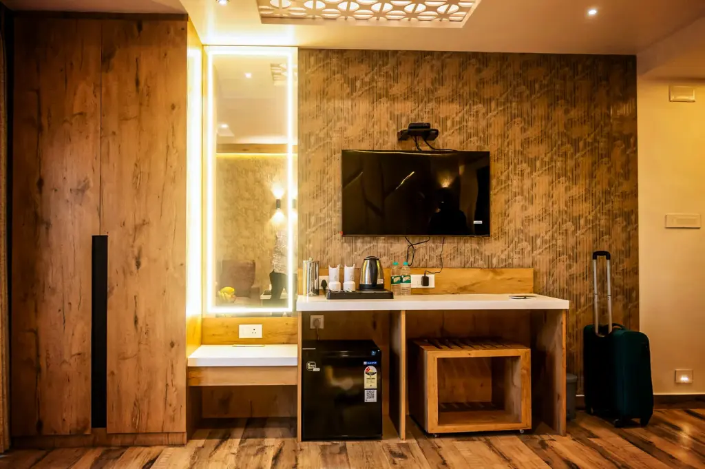 An eye-level shot captures the well-appointed amenities wall of a hotel room, representing the file "2-Why the Right Hotel Can Change.webp". On the left, a tall wardrobe with a textured light-wood finish stands next to a built-in vanity station, which features a long, vertically oriented mirror illuminated by a bright, continuous white border light. To the right, a flat-screen television is mounted on a textured, gold-and-beige patterned wall above a white-topped desk counter. The counter is equipped with a coffee station hosting a stainless steel kettle, mugs, and bottled water, while a mini-fridge and a wooden luggage rack are neatly tucked into the compartments below. On the far right, a dark teal rolling suitcase stands upright on the warm, multi-toned hardwood floor, completing the ready-for-travel atmosphere.