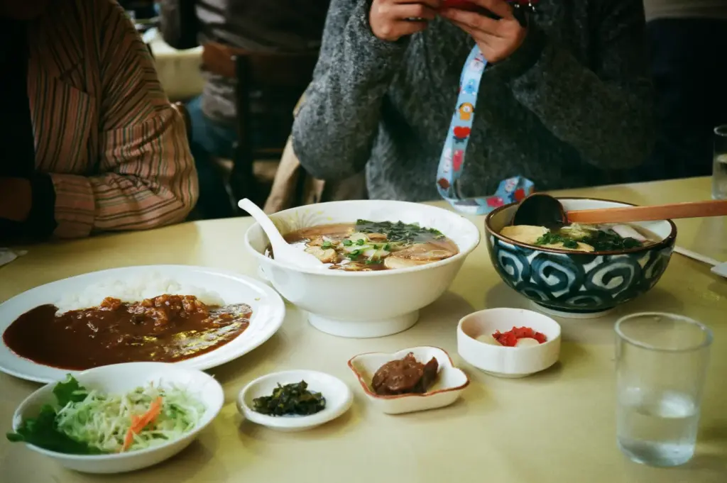 An eye-level, medium shot captures a casual restaurant table filled with a variety of traditional Japanese dishes, representing the file "3-Soup Curry The Bowl.webp". In the center stands a large, white porcelain bowl overflowing with hot ramen in a dark broth, topped with sliced meat, green onions, and nori seaweed, with a white soup spoon resting inside. To its right is a patterned blue-and-white ceramic bowl containing a separate noodle soup with a wooden serving ladle, while a white plate on the left holds a serving of rich Japanese curry alongside white rice. The meal is completed by small side dishes arranged across the foreground, including shredded cabbage salad, pickled condiments, and a glass of water, with two diners partially visible in the background as one holds a phone above the table.