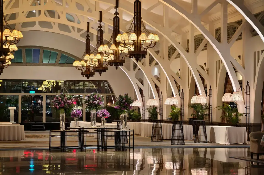 An eye-level, wide shot captures the expansive and architectural interior of a luxurious event hall, representing the file "3-The Fullerton Bay Hotel.webp". The space is defined by a massive, vaulted white ceiling supported by a striking row of white, geometric arched pillars lining the right wall, with each arch illuminated by its own slender, floor-standing lamp with a pleated shade. A long row of heavy, multi-tiered brass chandeliers with individual pleated shades hangs down the center, casting a warm, golden glow over the room. Below, several tables covered in white tablecloths are set up alongside tall, black wire cocktail tables, while the center stage features black console tables adorned with towering, vibrant purple flower arrangements. The highly polished dark marble flooring in the foreground reflects the warm lights and colorful floral displays, enhancing the grand, sophisticated atmosphere.