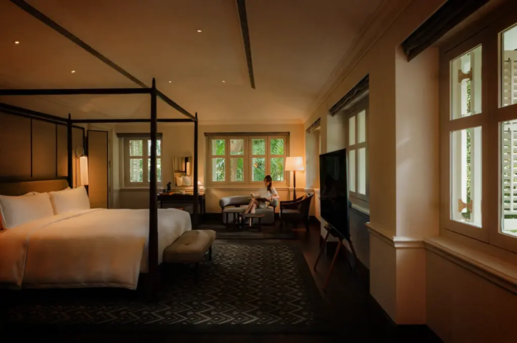 An eye-level, wide shot captures a spacious, elegantly appointed luxury hotel suite, representing the file "3-Where Stillness Meets Luxury.webp". In the foreground on the left, a large four-poster king bed is neatly made with crisp white linens, flanked by a padded bench at its foot and set over a dark, geometric patterned area rug. In the background, a woman in a white dress sits comfortably on a grey chaise lounge near a cluster of paneled windows that look out onto lush green foliage. A tall floor lamp casts a warm glow over a nearby armchair, while a modern flat-screen television stands on an easel to the right, all tied together by soft, recessed ceiling lighting that accentuates the room's calm, sophisticated atmosphere.