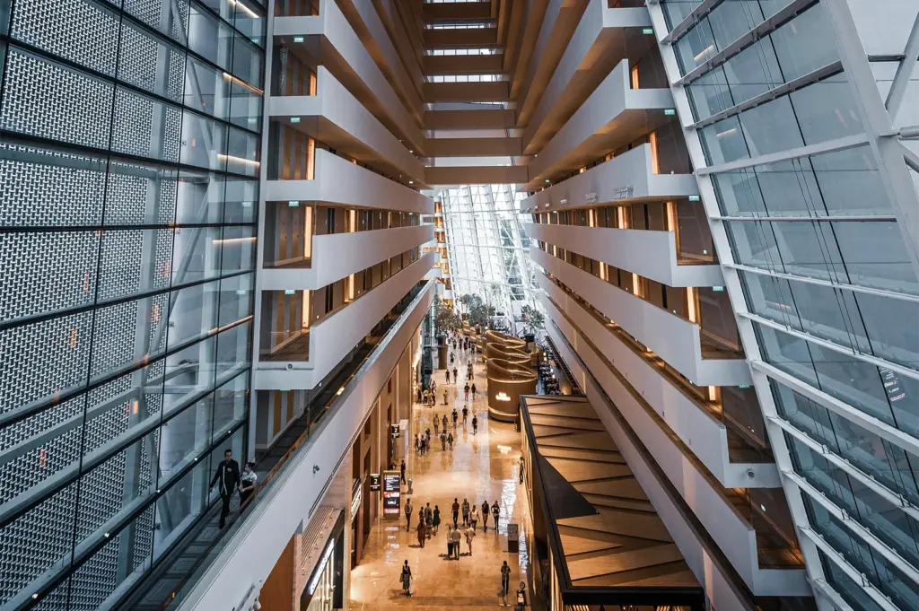 A high-angle, symmetrical perspective shot looks down into the massive, futuristic atrium lobby of a luxury hotel, representing the file "3-Why the Right Hotel Can Change_2.webp". Towering on both sides are interior balconies of guest floors that stagger inward as they ascend, creating a dramatic, geometric canyon-like view with warm light glowing from the corridor ceilings. The outer walls feature colossal, slanting glass and steel grid facades that flood the cavernous space with natural light. Far below on the polished, reflective marble ground floor, tiny figures of hotel guests stroll through the brightly lit concourse, passing by modern interior structures, indoor trees, and a low-slung restaurant pavilion on the right.