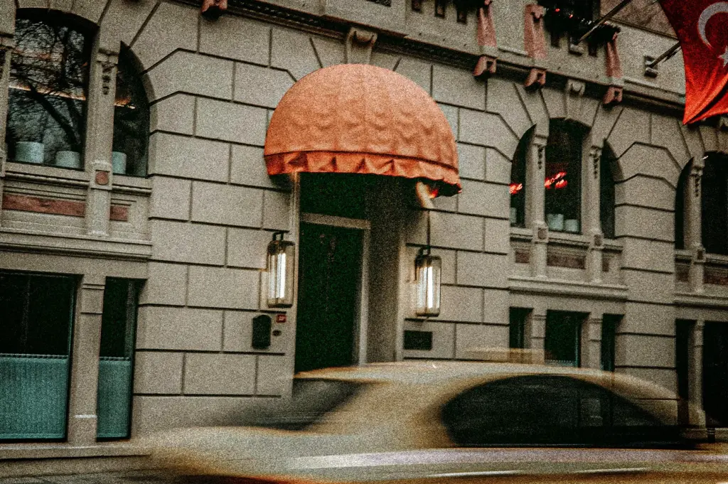 An eye-level, medium shot captures the classical stone facade of a building entrance, representing the file "4-Some Hotels Become Part.jpg". The entrance features a prominent, textured orange dome awning extending over a dark doorway, flanked by two glowing, vertical rectangular lanterns mounted to the light grey, rusticated stone blocks. Arched windows with ornate architectural moldings line the upper level, while a corner of a red Turkish flag is visible waving on the upper right. In the blurred foreground, the motion of a yellow taxi cab streaks past, creating a dynamic sense of urban movement across the historic and elegant architecture.