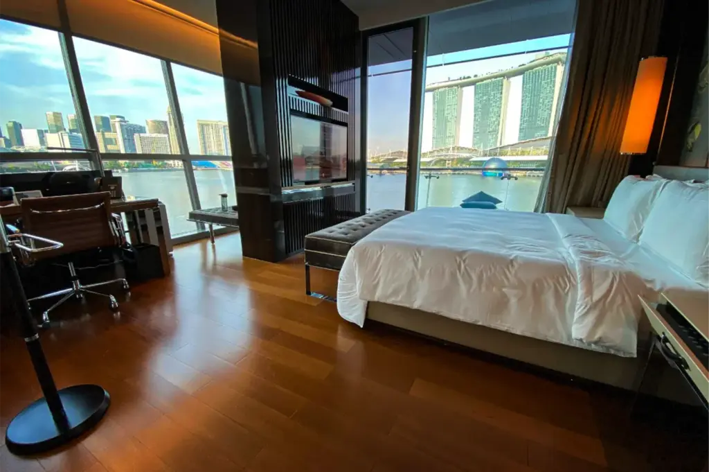 An eye-level, wide-angle shot captures a luxurious and modern hotel room with breathtaking waterfront views, representing the file "4-The Fullerton Bay Hotel.webp". On the right, a large bed with crisp white linens dominates the space, flanked by a tufted grey bench at its foot and a glowing floor lamp on the side. A dark wood-slat partition in the center houses a flat-screen television, dividing the sleeping quarters from a workspace on the left that features a brown leather rolling chair and a glass-top desk. Expansive, floor-to-ceiling glass windows and a sliding door reveal a scenic outdoor balcony overlooking a wide bay, where the iconic Marina Bay Sands resort and the distant city skyline stand prominently under a clear blue sky, casting bright, natural light across the room's polished hardwood flooring.