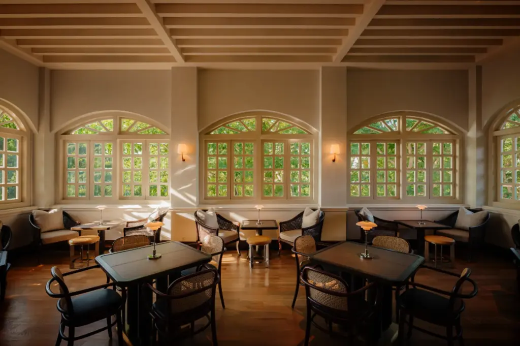 An eye-level, wide shot captures a bright, elegant resort lounge or dining area, representing the file "4-Where Stillness Meets Luxury.webp". The room features polished hardwood floors and a slatted beige ceiling, arranged with dark wood dining tables paired with cane-backed chairs in the foreground, each topped with a small, glowing table lamp. Lining the back wall are cozy seating alcoves with woven armchairs, plush cream-colored pillows, and small side tables, all situated beneath a row of large, arched multi-pane windows. Sunlight streams through the glass, casting soft shadows across the room and revealing a vibrant view of lush green trees outside, creating a serene and sophisticated colonial-inspired atmosphere.