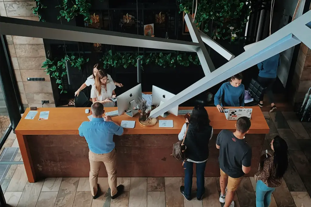 A high-angle, overhead shot captures a busy hotel reception counter on a tiled floor, representing the file "4-Why the Right Hotel Can Change.webp". Behind the long wooden desk, a staff member in a white t-shirt assists a guest with a payment terminal, another receptionist sits nearby, and a third employee in a blue sweatshirt works on a laptop covered in stickers. In front of the counter, a line of guests waits, including a man in a light blue shirt and tan pants at the far left, a woman with a dark vest and backpack in the middle, and a couple standing on the right. The modern, industrial-chic space is accented by sleek desktop computers, large abstract overhead light fixtures, and a black accent wall filled with lush green climbing plants in the background.