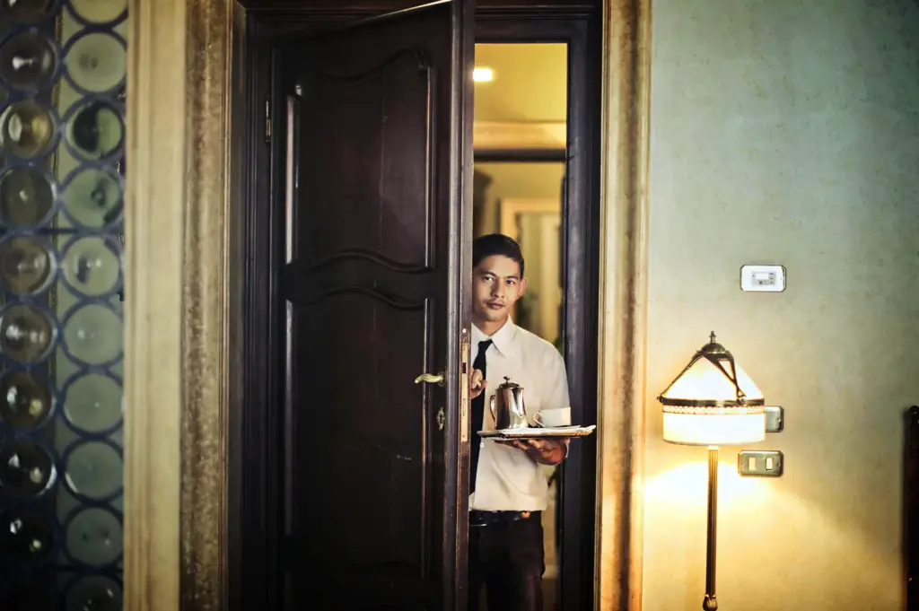 An eye-level, medium shot captures a hotel room service attendant stepping through a doorway, representing the file "5-Some Hotels Become Part.webp". The male attendant, dressed in a white button-down shirt and a black tie, looks directly toward the camera while holding open a large, ornate dark wood door with his right hand. In his left hand, he balances a silver serving tray containing a metallic coffee pot and a white ceramic teacup with a saucer. The softly lit room interior on the right features a textured beige wall, light switches, and a glowing traditional table lamp, creating an intimate atmosphere that highlights premium hospitality and personalized guest service.