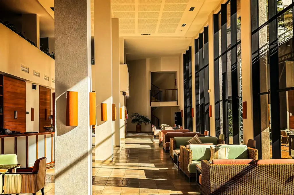 An eye-level, wide-angle shot captures the spacious, sun-drenched lobby of a hotel, representing the file "6-Some Hotels Become Part.webp". The architectural space is defined by an exceptionally high ceiling and a long row of massive, square concrete pillars on the left, each adorned with vertical orange cylindrical light fixtures that cast long shadows across the tiled flooring. To the right, a soaring wall of floor-to-ceiling glass windows floods the hallway with bright afternoon sunlight, illuminating a series of cozy seating arrangements made of wicker couches with pastel green and peach cushions. In the far background, a potted palm tree rests near a staircase landing, enhancing the relaxed, open, and airy resort atmosphere of the atrium.