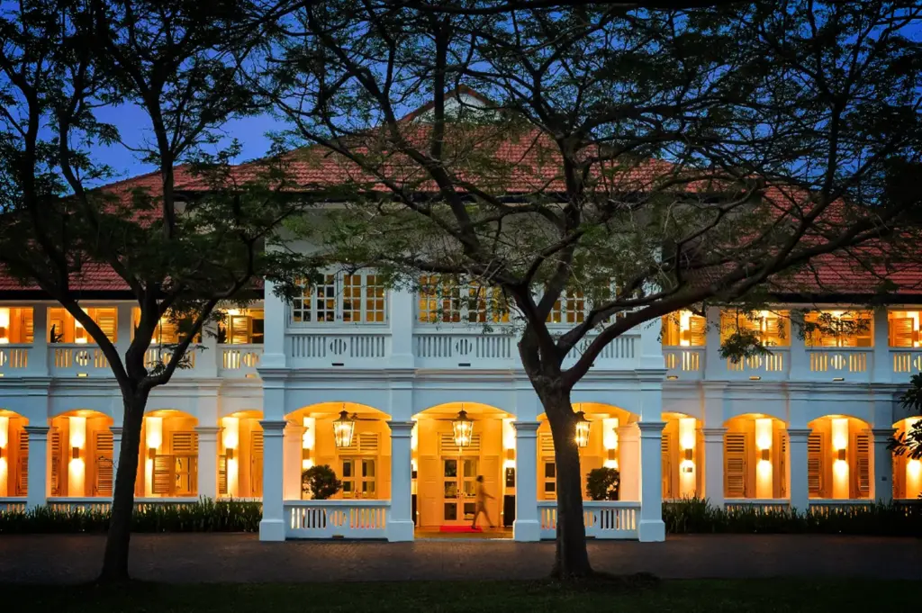 An eye-level, symmetrical shot captures the grand, illuminated exterior of a colonial-style white estate building during twilight, representing the file "6-Where Stillness Meets Luxury.webp". The two-story structure features a terracotta-tiled roof and rows of arched entryways and shuttered windows, all glowing warmly with bright interior and exterior lights. In the center foreground, a covered portico with hanging lanterns shelters a doorway where a blurred figure walks across a red carpet runner. Framing the symmetrical facade are two large, leafy trees whose dark branches stretch across the deep blue evening sky, adding a sense of historic charm, privacy, and quiet luxury to the scene.