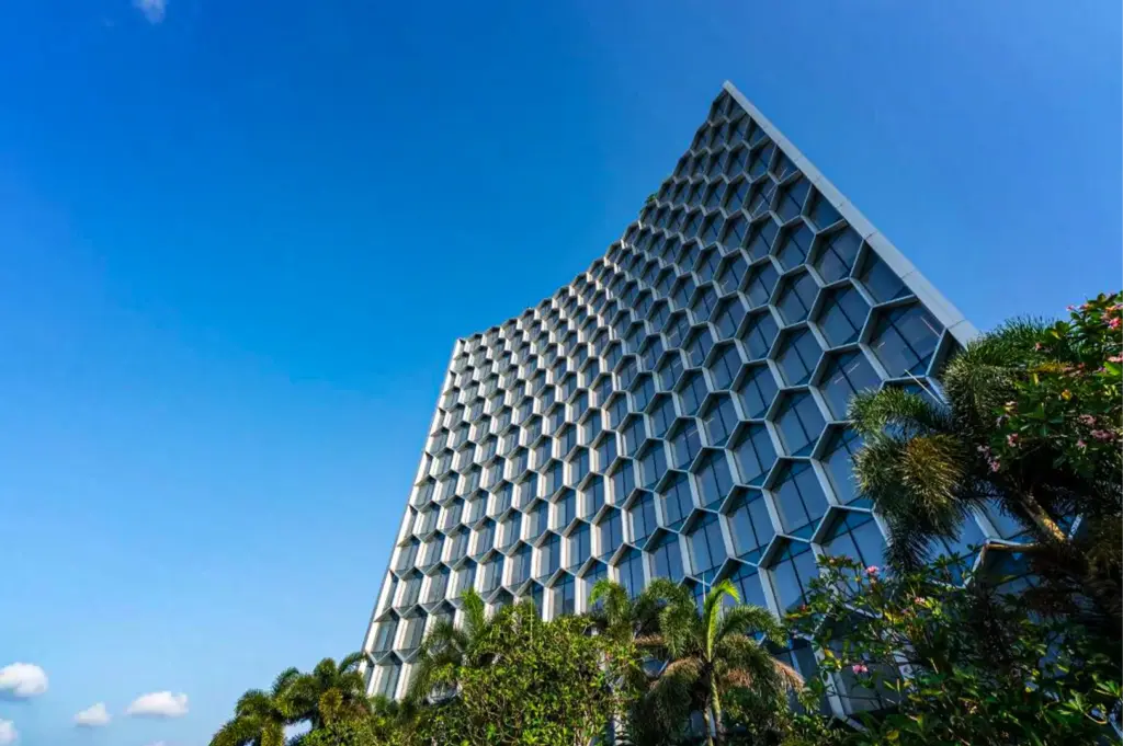 A low-angle, wide shot captures the striking modern facade of a high-rise tower against a clear, vibrant blue sky, representing the file "1-Andaz Singapore.webp". The building's exterior is defined by a unique, concave curved architectural design enveloped in a massive, repeating honeycomb matrix of hexagonal glass windows with light gray frames. Lush green palm trees and tropical shrubs line the bottom right foreground, partially obscuring the base of the structure, while the bright afternoon sunlight casts soft shadows across the geometric contours of the building's distinctive surface.
