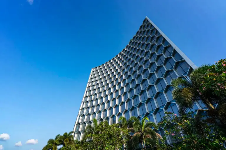 A low-angle, wide shot captures the striking modern facade of a high-rise tower against a clear, vibrant blue sky, representing the file "1-Andaz Singapore.webp". The building's exterior is defined by a unique, concave curved architectural design enveloped in a massive, repeating honeycomb matrix of hexagonal glass windows with light gray frames. Lush green palm trees and tropical shrubs line the bottom right foreground, partially obscuring the base of the structure, while the bright afternoon sunlight casts soft shadows across the geometric contours of the building's distinctive surface.