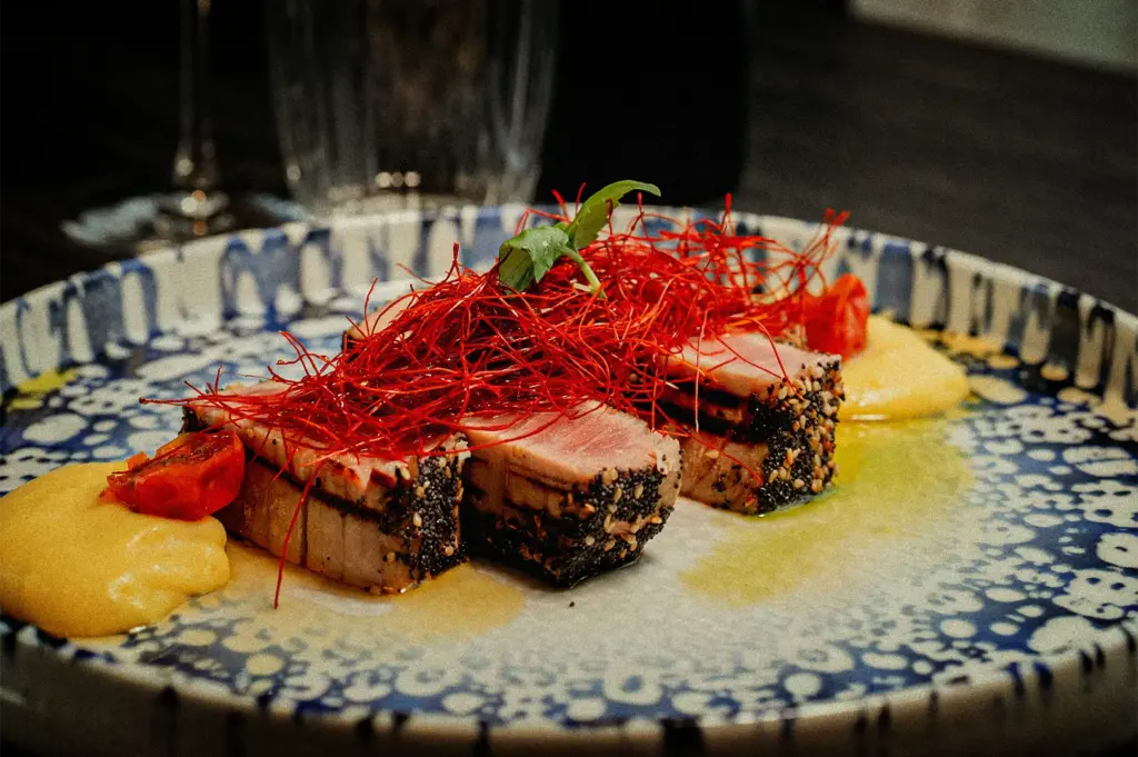 A close-up, slightly high-angle shot captures a beautifully plated seafood dish presented on an artisanal blue and white patterned ceramic plate, representing the file "1-Every Great Tasting Menu.jpg". Several thick slices of seared tuna, cooked rare with a pink center and crusted in black poppy seeds and sesame seeds, are arranged neatly in the center of the plate. The tuna is topped with a vibrant, tangled nest of fine, bright red chili threads and a single green microgreen leaf. Creamy dollops of a yellow puree or sauce and small roasted cherry tomato halves flank the fish, all drizzled with a light sheen of olive oil, while a blurred wine glass sits in the dark background.