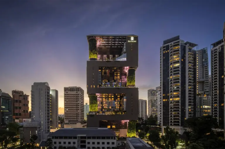 A wide-angle, eye-level cityscape shot captures a stunning modern skyscraper at dusk, representing the file "1-Inside Pan Pacific Orchard Luxury.webp". The building features an innovative, block-like geometric architectural design with massive, open-air cutouts that contain multi-story vertical gardens, illuminated in a vibrant green glow. Warm interior lights peek through the windows of the hotel rooms, and the illuminated "PAN PACIFIC" logo is clearly visible near the top-right corner. Flanked by other high-rise apartments and office buildings, the structure stands out majestically against a transitioning twilight sky that fades from a warm orange sunset glow on the left to a deep blue-purple overhead.