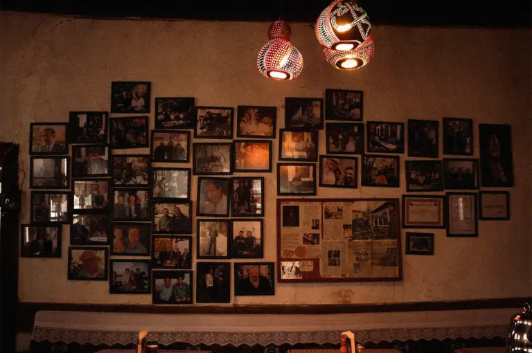 A warmly lit restaurant wall decorated with an extensive gallery of framed photographs, certificates, newspaper clippings, and memorabilia that showcase the establishment’s history and notable visitors. Colorful handcrafted pendant lights hang from the ceiling, casting a cozy glow over the display and creating a nostalgic atmosphere. The collection of personal photographs and awards adds character to the dining space, reflecting the restaurant’s heritage, community connections, and long-standing reputation.