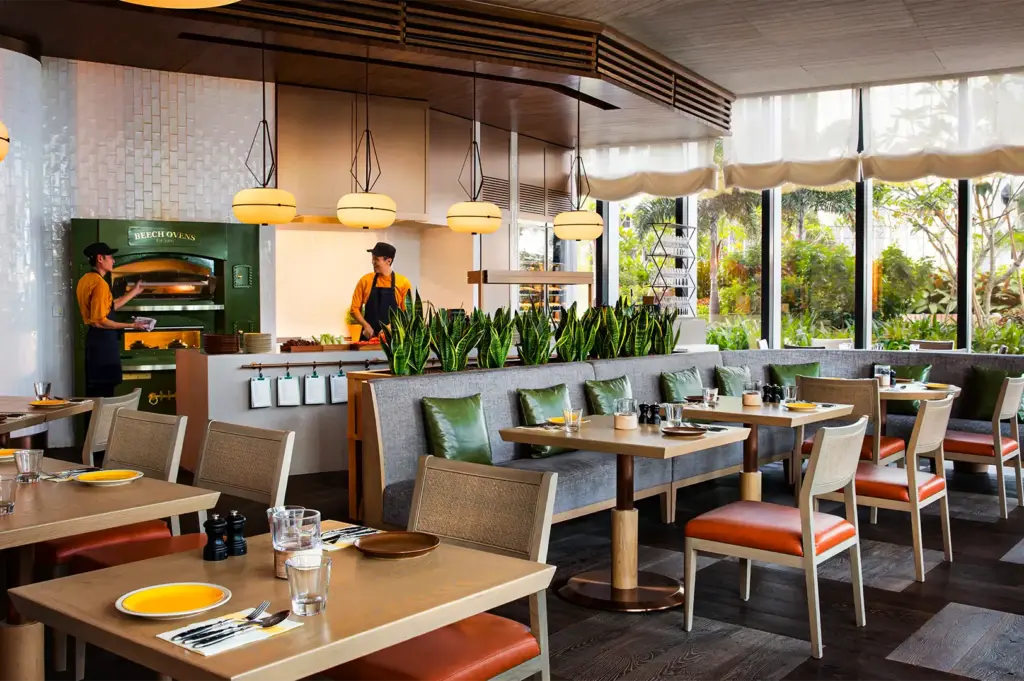 A wide-angle, eye-level shot captures a bright and modern hotel restaurant dining room with an open show kitchen, representing the file "3-Andaz Singapore.webp". In the background, two chefs in yellow uniforms and dark aprons work near a large green "BEECH OVENS" station and a food preparation counter topped with a row of vibrant snake plants. The dining area is furnished with light wood tables set with yellow and brown plates, surrounded by modern chairs with orange leather seats and a long gray banquette sofa accented by green leather pillows. Four large, oval pendant lights hang from a wooden ceiling feature, while expansive floor-to-ceiling windows on the right flood the room with natural light and look out onto lush tropical greenery.