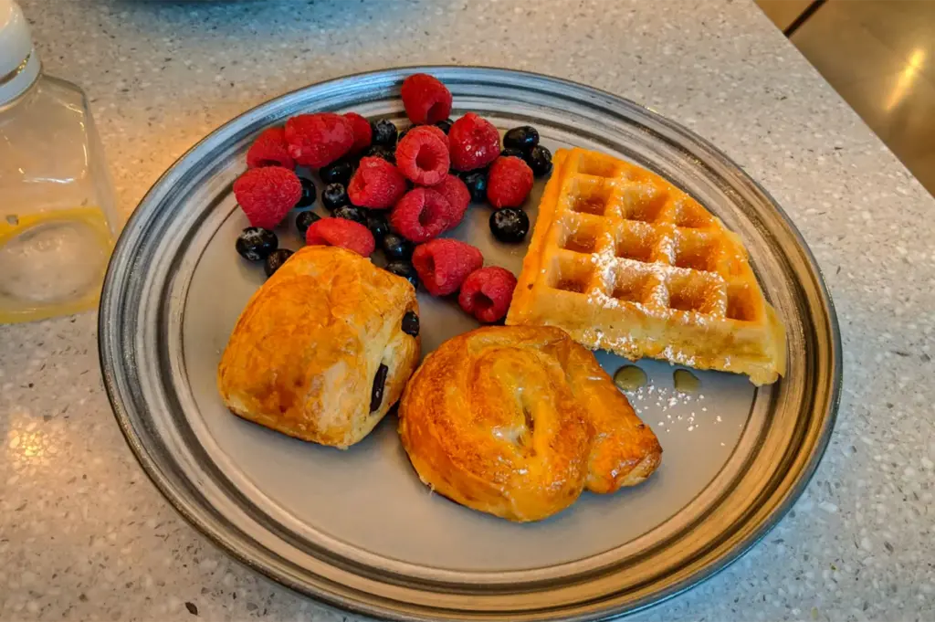 A high-angle, close-up shot captures a breakfast plate filled with pastries and fresh berries on a speckled gray countertop, representing the file "4-Andaz Singapore.webp". Arranged on a round ceramic plate with gray concentric rings, the meal features a golden, triangular waffle section dusted with powdered sugar and drizzled with syrup, a flaky pain au chocolat, and a swirled Danish pastry. A generous pile of vibrant red raspberries and dark blueberries occupies the top section of the plate, while a glass bottle containing a yellow liquid is partially visible in the upper-left corner.